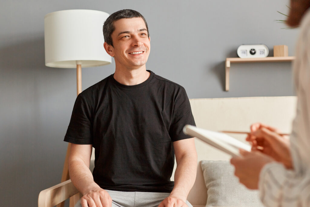 Inspired handsome young man in black T-shirt sitting on couch at counselor office, sharing feelings, thoughts with therapist woman, enjoying therapy session.