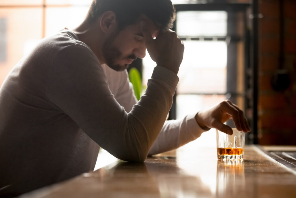 Young stressed drunk man sitting alone with glass of whiskey in bar in the morning, thinking about problem at work or relationships, feeling lonely and unhappy, alcohol addiction concept